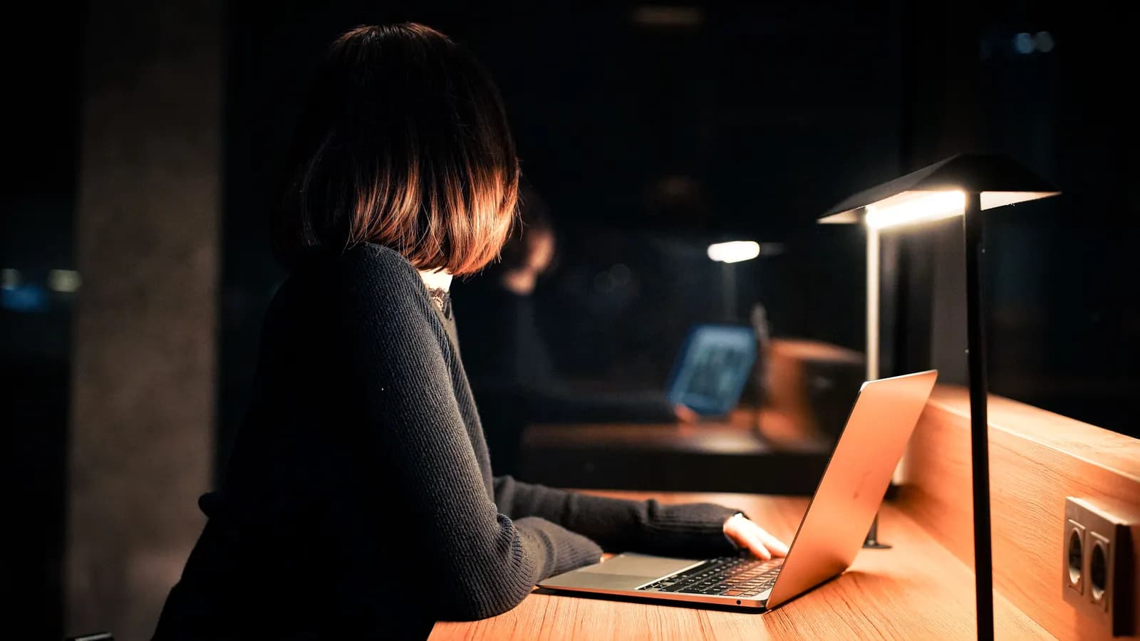 Une personne travaille tard le soir sous une lampe de bureau, gardant un œil sur son écran